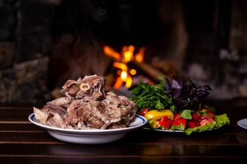 Against the backdrop of a fireplace with crackling logs, there is a plate of steaming boiled meat and a bowl of fresh vegetable salad.