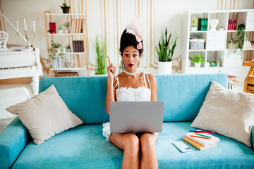 Elegant young woman in historic attire using a laptop on a modern couch surrounded by luxurious...