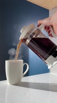 Pouring freshly brewed coffee from a French press into a white mug, with steam rising gracefully in a cozy kitchen setting against a dark blue wall. A perfect moment for a coffee break