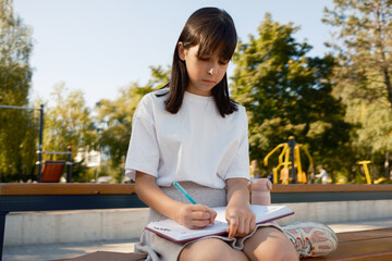 A young girl concentrates as she writes in her notebook while sitting on a bench outdoors....