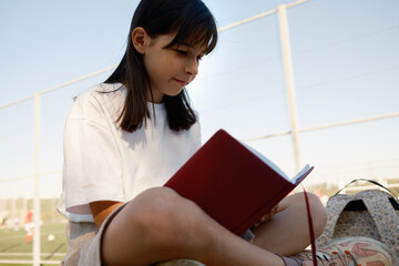 A young girl sits outdoors in a park, engrossed in reading a book. The sunny environment and casual...