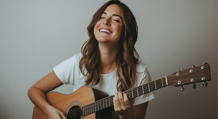 Fototapeta premium smiling woman with wavy brown hair plays an acoustic guitar, radiating a relaxed and joyful mood. She wears a simple white shirt, creating a casual, warm atmosphere.