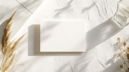 Wedding RSVP card mockup placed on a textured white surface with soft natural lighting. Minimalist design with bright white paper, neutral accents like glass, a white tile, and delicate dried grass.