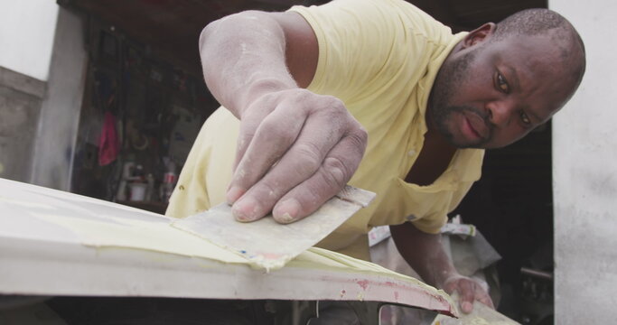 Front view of an African male panel beater in a township workshop, painting body of a car, slow moti