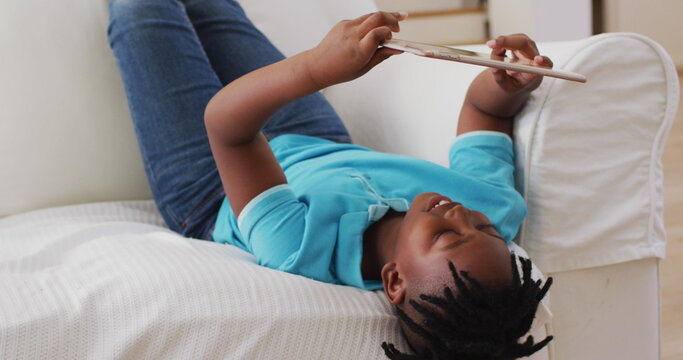 African american boy smiling while using digital tablet while lying on the couch at home
