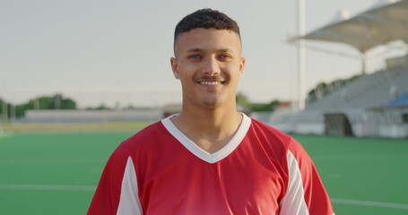 Portrait close up of a biracial male field hockey player, wearing a red team strip, standing on a ho