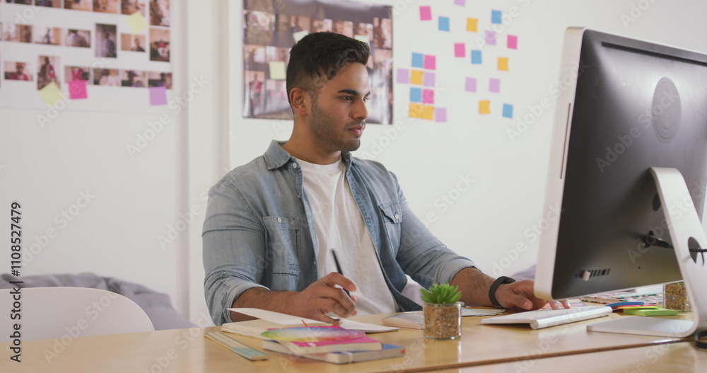 Poster front view of a biracial man working in a creative office, looking at computer screen and writing at