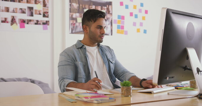 Front view of a biracial man working in a creative office, looking at computer screen and writing at