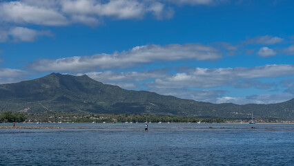 View of a tropical island from the ocean. A mountain against the blue sky and clouds. A city on the...