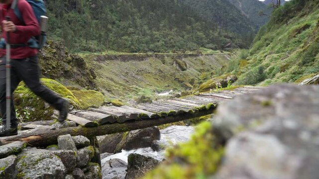 Man with backpack and trekking poles crossing mountain creek wooden bridge during Makalu Barun National Park trek in Nepal. Mountain hiking and active people concept image.