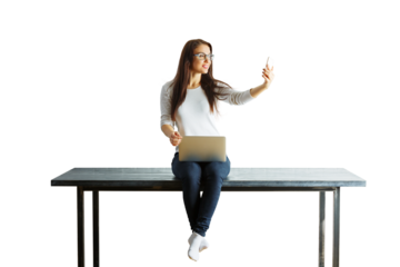 Woman sitting on a table, holding a laptop and taking a selfie with her phone. Isolated on a white background. Concept of multitasking and technology