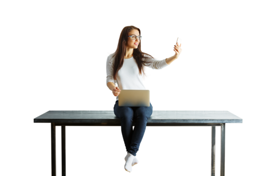 Woman sitting on a table, holding a laptop and taking a selfie with her phone. Isolated on a white background. Concept of multitasking and technology - Powered by Adobe