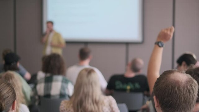 Audience member raising hand to ask a question during a business presentation. Engaging speaker discussing key topics with group of diverse attendees in a conference setting.