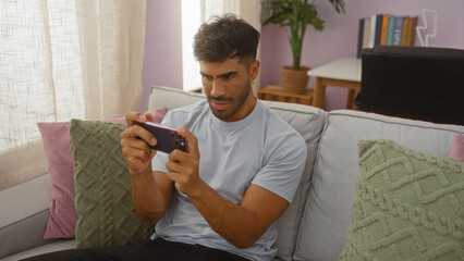 Young hispanic man playing on smartphone in cozy living room with pastel decor and plush cushions