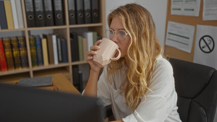 Young woman with blonde hair in office environment drinking coffee while sitting at desk,...