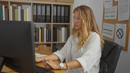 Young blonde woman working intently at a computer in a well-organized office filled with shelves of...