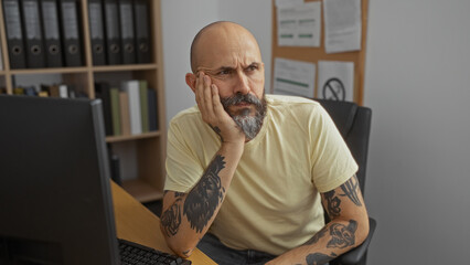 Hispanic man with beard and tattoos sits at office desk indoors, deep in thought, surrounded by...