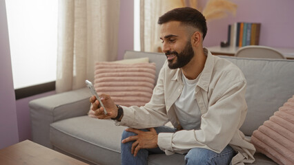 Young hispanic man sitting in a cozy living room using a smartphone, wearing casual clothes and smiling while relaxed at home.