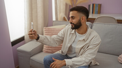 Fototapeta premium Young man taking a selfie in a cozy living room with contemporary decor, featuring beige couches and sunlight streaming through the window