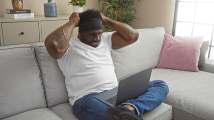 A young african american man sitting on a sofa in his living room, frustrated while using a laptop, surrounded by cozy home decor and indoor plants.