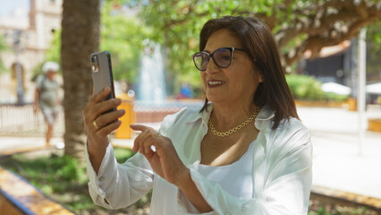 Mature hispanic woman with short hair uses a smartphone while smiling outdoors in a sunny urban park.