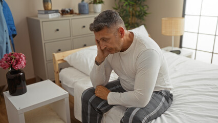 Fototapeta premium Middle-age man sitting on a bed in a bedroom looking thoughtful, indoors, wearing pajamas and a white shirt, surrounded by simple interior decor including flowers, a lamp, and dresser.