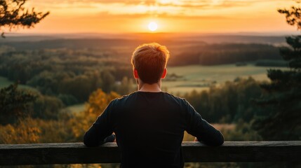 Fototapeta premium Young man enjoying a serene sunset view overlooking an expansive landscape with trees and fields, capturing a moment of tranquility and reflection in nature.