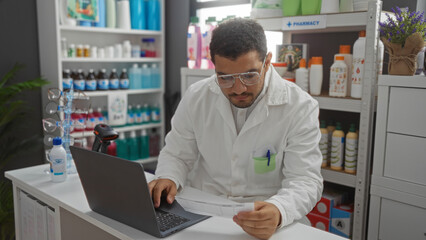 Young hispanic man working at a laptop in a pharmacy or drugstore, checking a prescription in a well-organized store interior