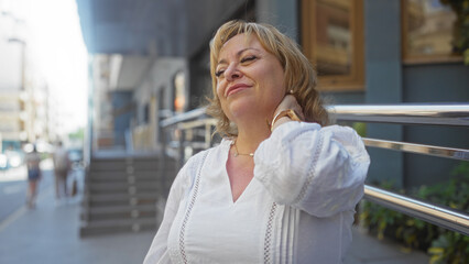 Fototapeta premium Middle-aged woman in a white blouse holding her neck in discomfort on an urban street with buildings and people in the background.
