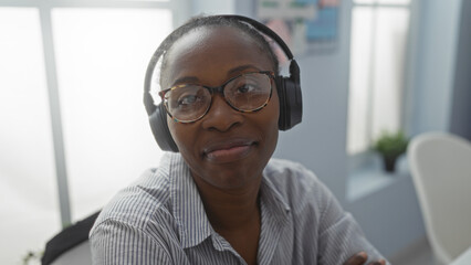 Fototapeta premium Woman wearing headphones and glasses, seated in an office with a striped shirt, looking content against a bright indoor background