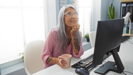 Woman working in an office, looking thoughtful with a cup of coffee while sitting at a desk with a...