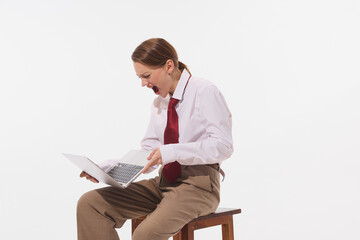 Young woman in white shirt and red tie sitting on wooden stool, yelling at her laptop in frustration against white studio background. Concept of business, leadership, occupation, modern culture. Ad