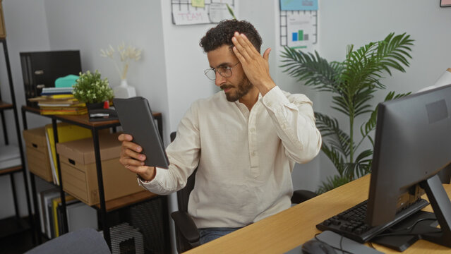 Handsome young man in office peines hair with hand while looking at tablet, surrounded by indoor plants and documents on desk.
