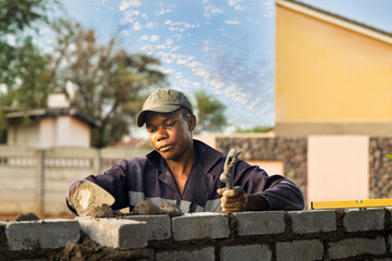 african bricklayer with a trowel , spirit level and a hammer building a surrounding bricks wall,...