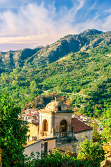 View of italian town in mountain valley of Sicily, Italy. Old town in a valley among green mountains with ancient church and vintage roofs of buildings in beautiful evening sunset light