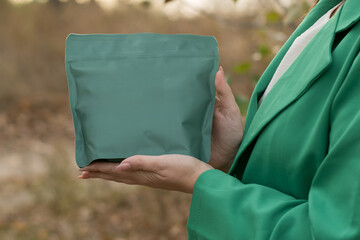 Person in a green coat presenting a green package outdoors, surrounded by natural greenery, with clear space for text or design elements. Suitable for marketing eco-friendly products  