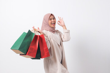 Portrait of excited Asian hijab woman in casual suit holding paper shopping bags while making ok hand gesture. Shopaholic girl and discount buying concept. Isolated image on white background