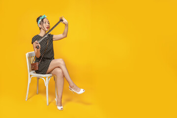 Fototapeta premium Asian girl dressed in pin-up style with vintage camera emotionally looking at developed film negative on bright yellow background. Isolated