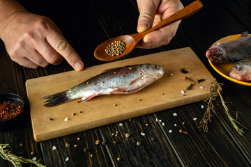 A skilled cook prepares a fresh fish on a wooden cutting board, carefully sprinkling spices with...