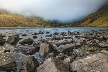 Smooth lake waters reflecting the overcast sky, with large rocks scattered along the lakeshore.