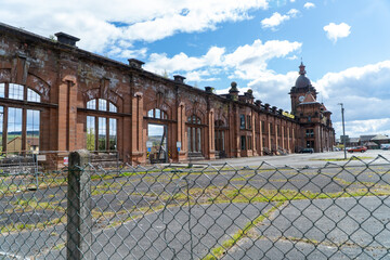 A vacant parking lot fronts a decayed red-brick facade with arched windows, framed by a wire fence. The building’s timeless architecture contrasts with its weathered state under a bright blue sky.