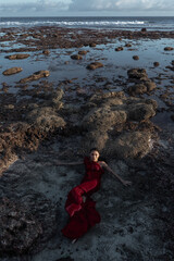 Beautiful girl submerged in water in a red dress on a reef near the ocean