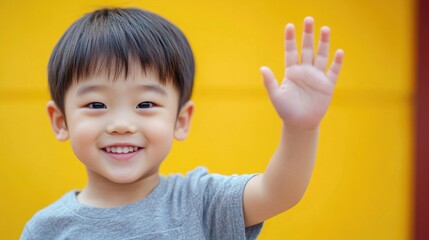 A young boy raises his hand in front of a bright yellow wall, a simple yet cheerful scene