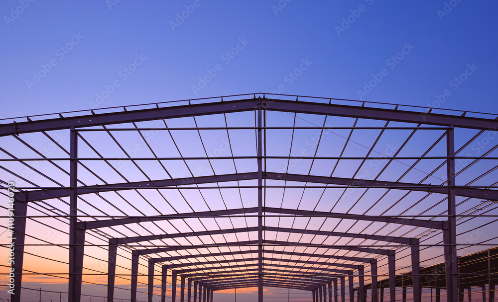 Wall mural Silhouette of large factory building framework with metal roof beam and post structure in construction site against evening sky background,  perspective front view