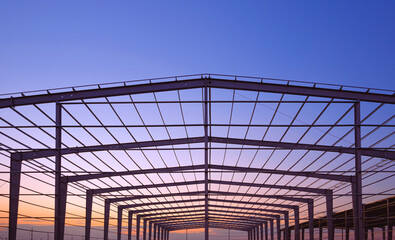 Silhouette of large factory building framework with metal roof beam and post structure in construction site against evening sky background,  perspective front view