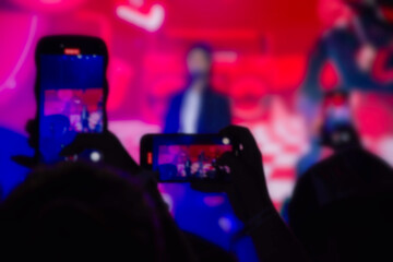 Fans in an audience on a concert recording on their smartphones. Red and blue background.