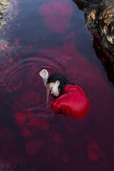 Beautiful girl submerged in water in a red dress on a reef near the ocean