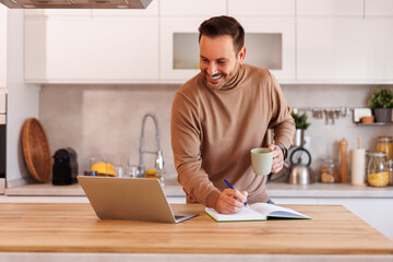 Mid adult man holding coffee mug and writing in note pad while working over laptop at table in...