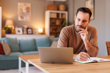 Portrait of focused businessman writing in diary while doing online research over computer in home...
