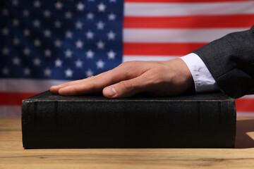 Man taking oath with his hand on Bible at wooden table against flag of USA, closeup